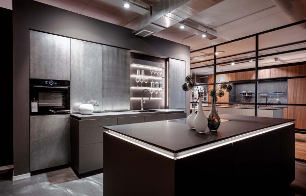 Minimalistische Designer-Küche mit Betonoptik-Fronten, beleuchteten Glasregalen und schwarzer Kücheninsel bei Gellert Küchenwelten.