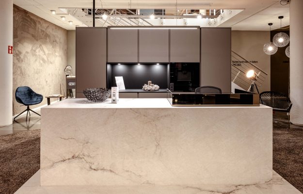 Premium-Inselküche mit heller Steinverkleidung, minimalistischen Fronten und stimmungsvoller Beleuchtung – luxuriöses Ausstellungskonzept bei Gellert Küchenwelten.